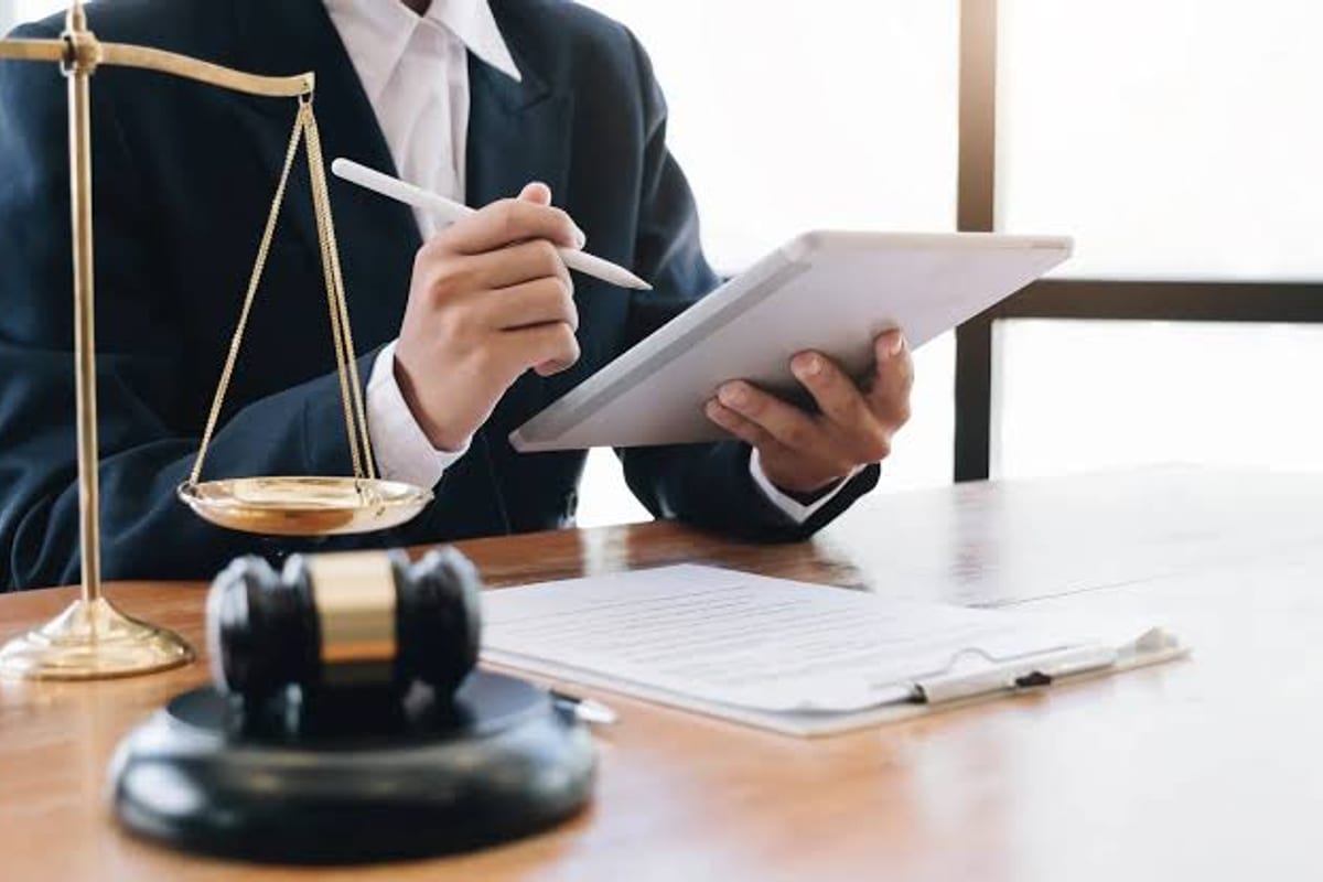 A desk with a set of brass scales, a gavel and a document, someone reading from a tablet above them.