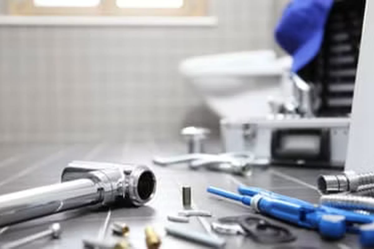 A bathroom floor covered in plumbing parts — taps, hoses, pipe sections and wrenches — with someone working at the toilet behind.