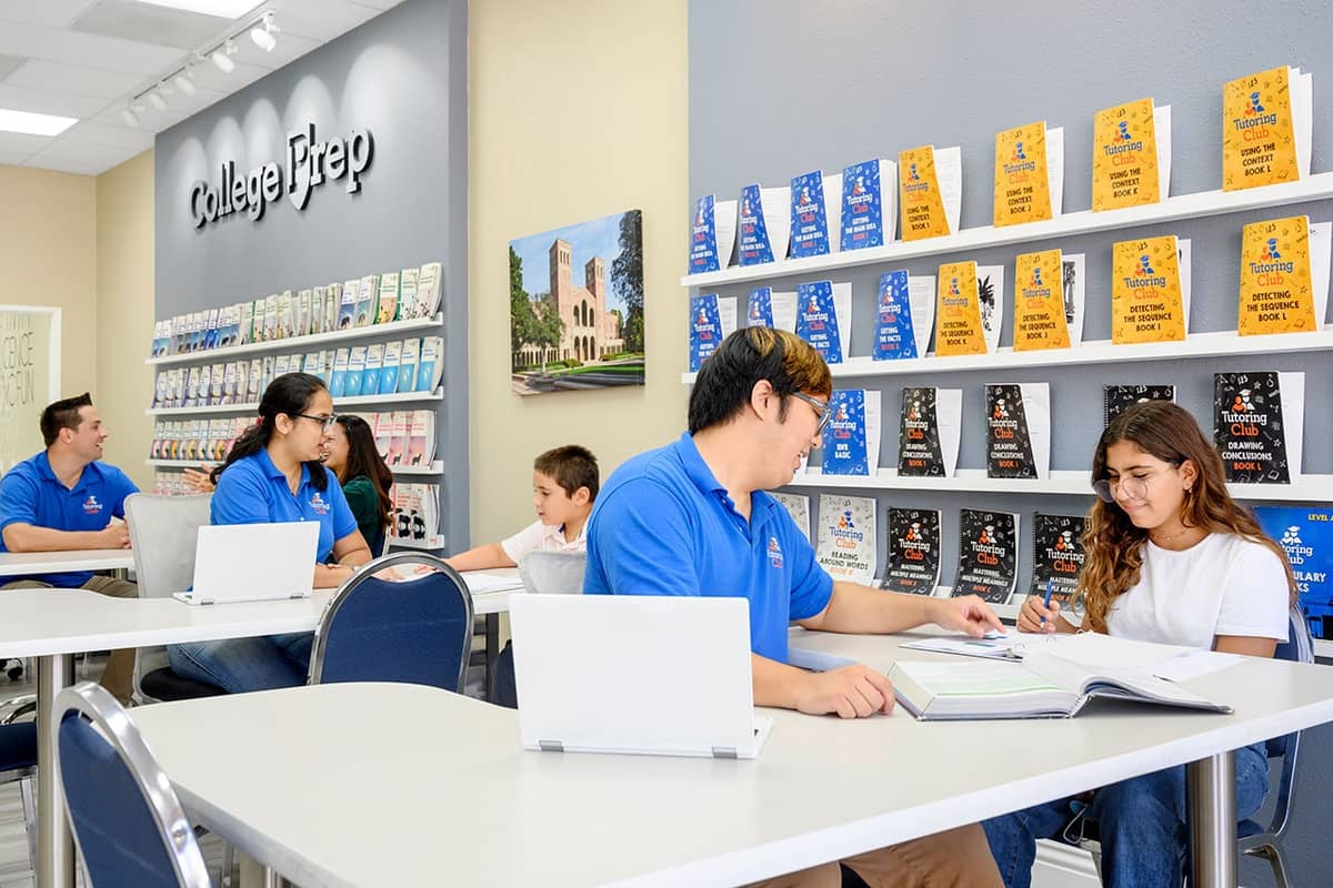 A tutoring centre: tutors working one to one with students at white tables, workbooks on the shelves behind.